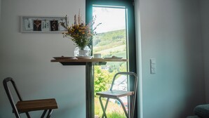 A room with a window, a chair, a table with a vase of flowers, and a wall-mounted shelf with pictures. - bikehikesleep (Sasbachwalden)