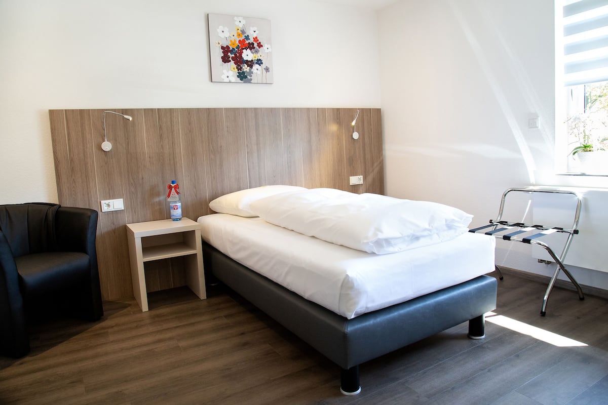 A modern hotel room with a wooden headboard, a bed with white linens, a small bedside table with a bottle, and a chair.
