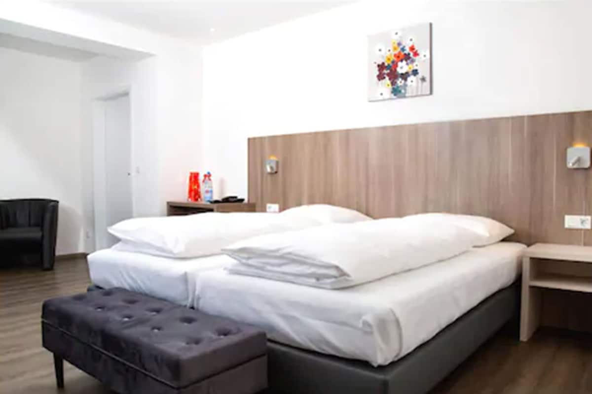 A modern hotel room with a large bed, a small bedside table, a nightstand, a grey ottoman, and a wall-mounted artwork.