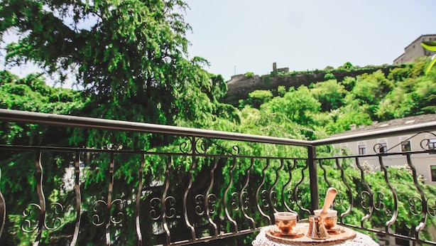 A balcony with a wrought iron railing, a small table with a decorative cloth, and a view of lush greenery and a building in the background. - Hotel RELAX Pazari i Vjeter (Gjirokastër)