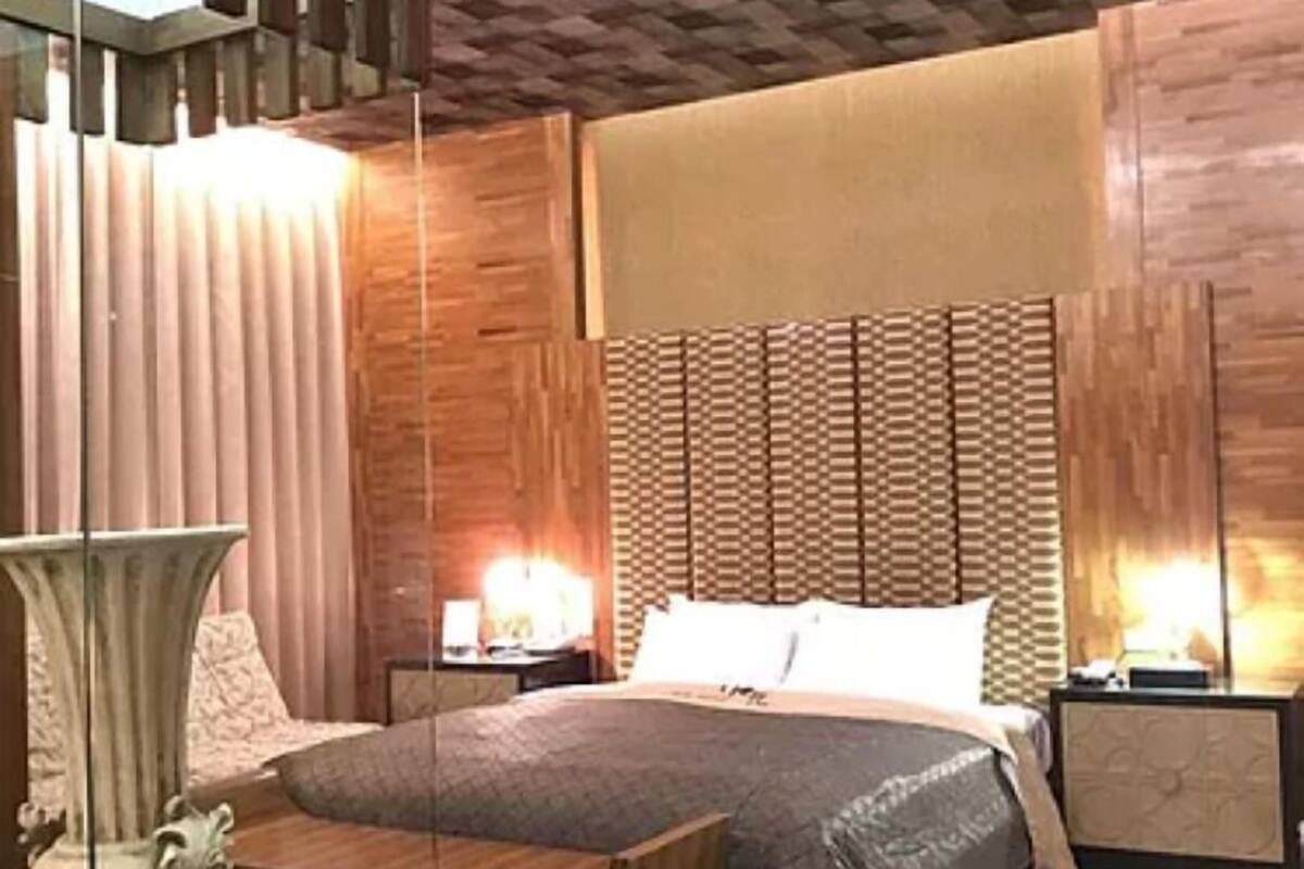 A modern bedroom with a wooden headboard, a bed with a grey quilt, a bedside table with a lamp, and a decorative vase.