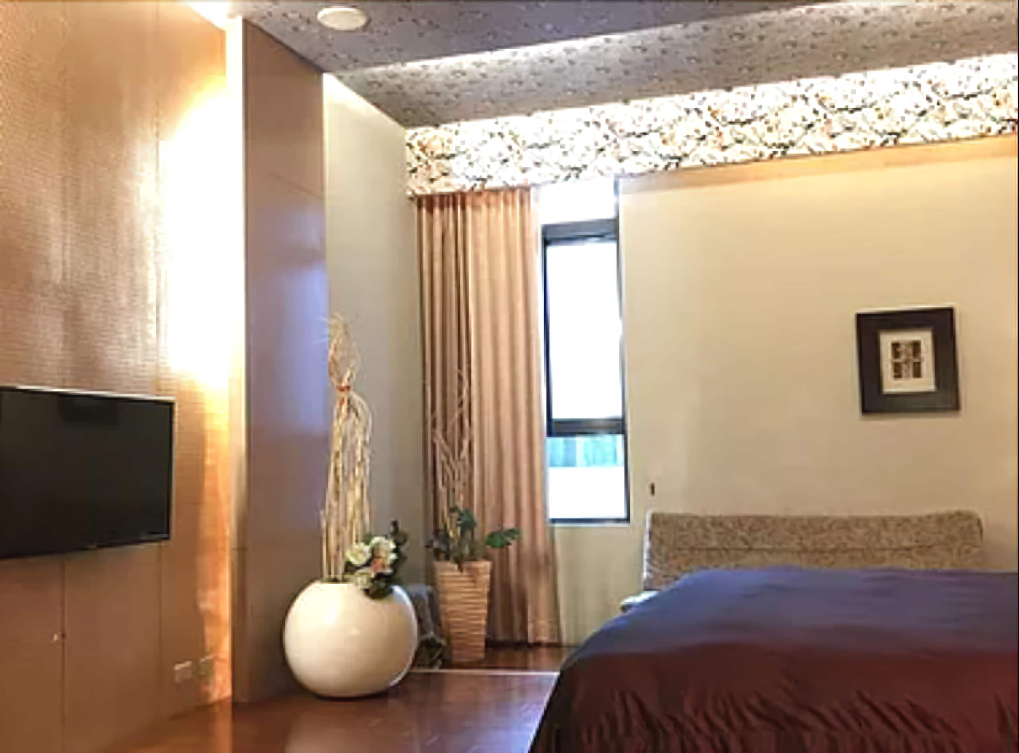 Una habitación de hotel con una cama, televisión, ventana con cortinas, un cuadro en la pared y un jarrón con flores.