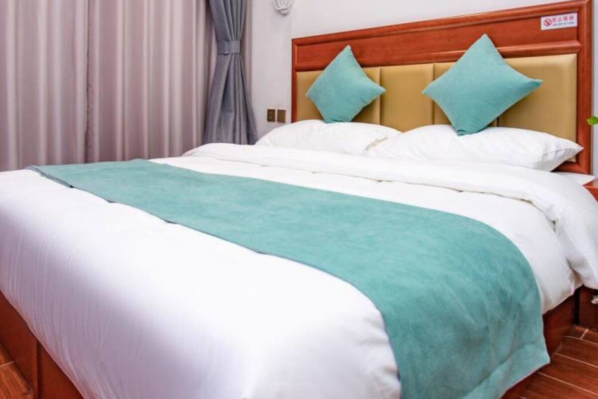 Una cama bien hecha con una colcha azul turquesa y ropa blanca, un cabecero de madera con dos almohadas azul turquesa y una cortina gris a la izquierda.