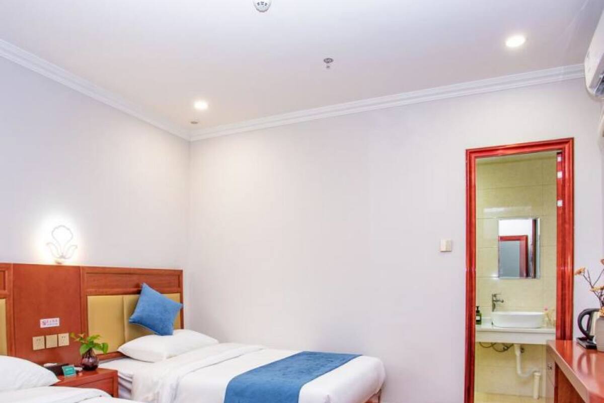 Habitación de hotel con dos camas, un cabecero de madera, una mesita de noche con lámpara y un baño visible a través de una puerta de vidrio.