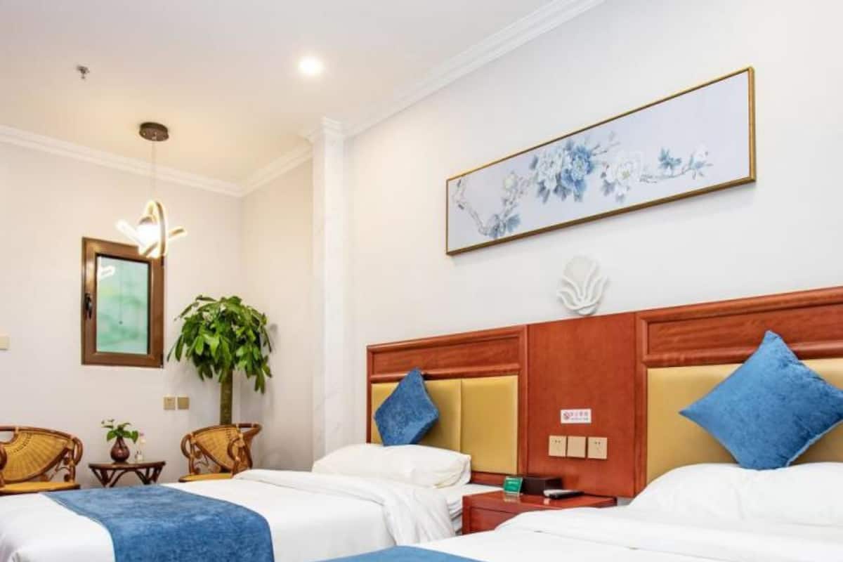 Habitación de hotel con dos camas, cabecero de madera, un cuadro grande con motivos florales, una planta en maceta y una mesita con lámpara.