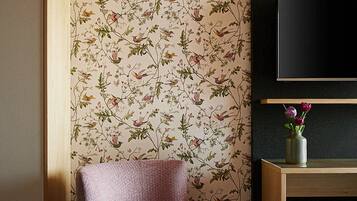 Une pièce avec un papier peint à motifs floraux, une chaise rose, un lit, un téléviseur fixé au mur et un petit vase avec des fleurs sur une étagère en bois.