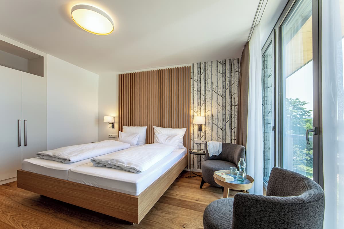 Una habitación de hotel moderna con una cama grande, un área de descanso con una pequeña mesa y vistas al exterior a través de amplias ventanas.