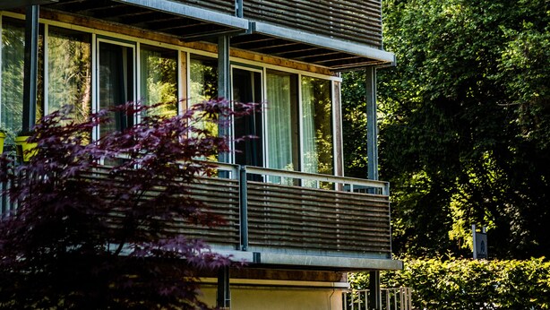 A multi-story building with balconies and large windows, surrounded by greenery.