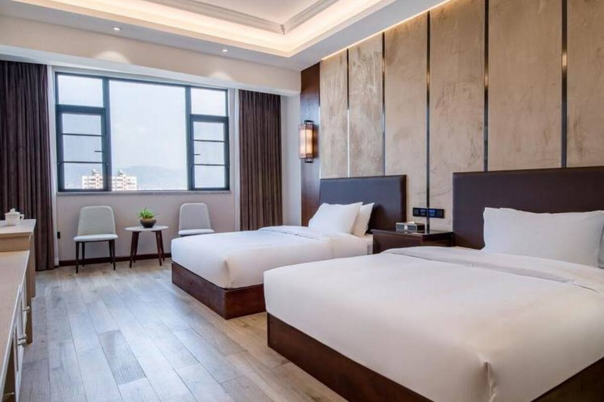 Habitación de hotel con dos camas, un escritorio, una silla, una mesita y una ventana con vistas a edificios.