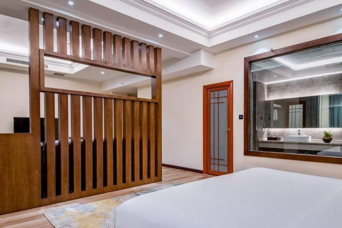 Un dormitorio moderno con una cama grande, un separador de madera, un espejo amplio y un baño visible a través de la puerta de cristal.