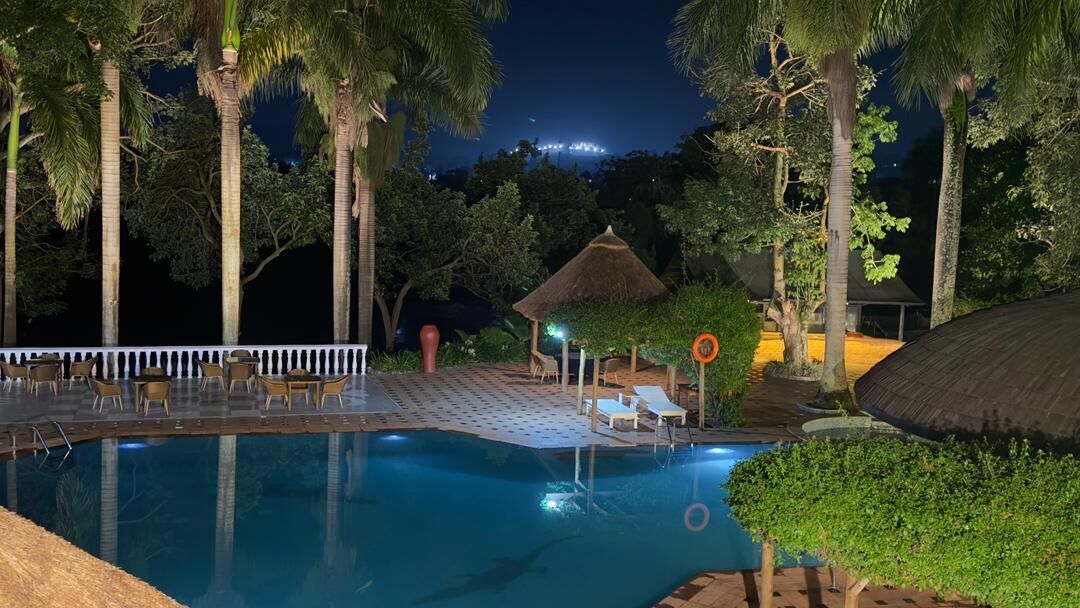 A nighttime pool area with lounge chairs, a thatched-roof structure, and surrounding trees.