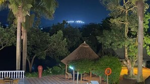 Un área de piscina nocturna con sillones reclinables, una estructura de techo de paja y palmeras.