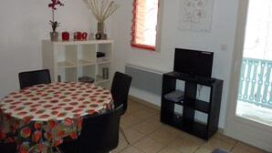 A dining area with a round table covered in a strawberry pattern, surrounded by chairs. There is a white shelving unit with decorative items and a television on a stand.