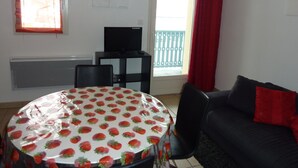 A room with a round table covered in a strawberry-patterned tablecloth, a black sofa with red cushions, a television on a black stand, and a window with a red curtain.