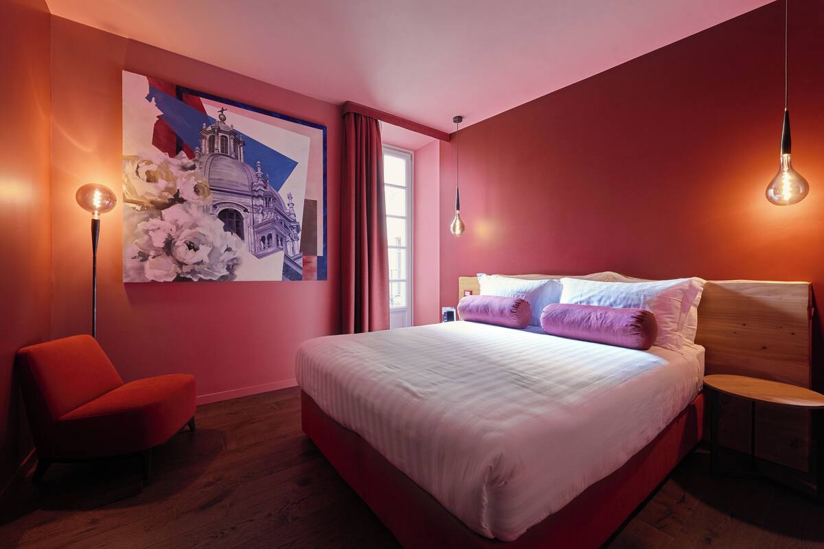 A bedroom with a large bed, a red chair, a wooden nightstand, and a wall painting.