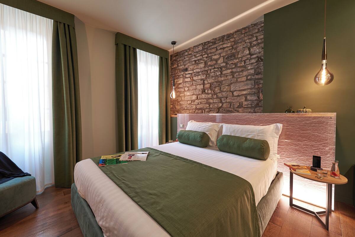 A modern hotel room with a large bed, green curtains, a stone wall, and a small table with a lamp.
