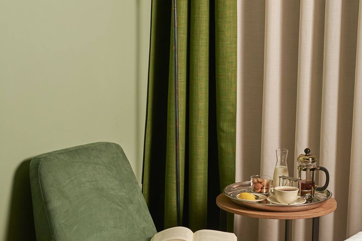 A green armchair with an open book, a bedside table with a coffee pot, a glass of milk, and a tray with food and a cup.