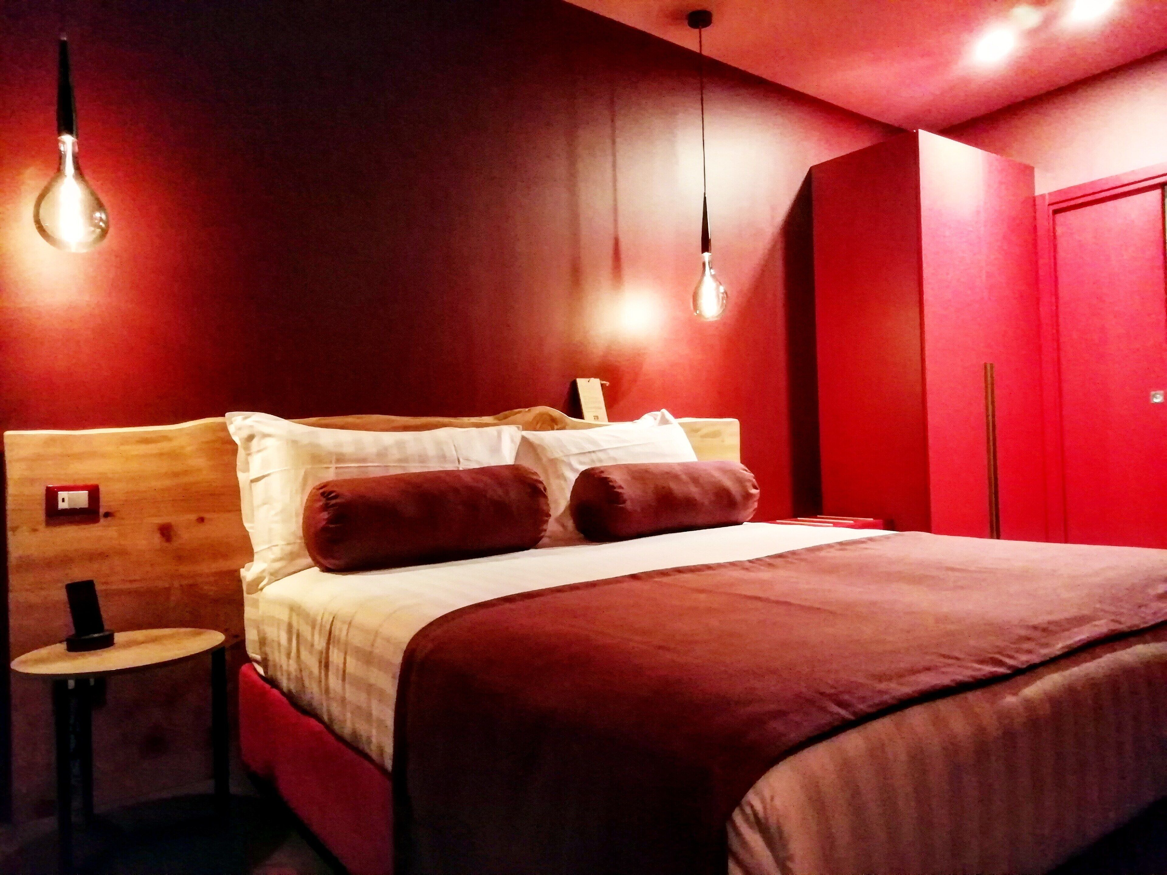 Quarto de hotel com cabeceira de madeira, cama com duas almofadas, mesa de cabeceira com luminária e guarda-roupa vermelho.