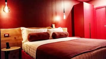 Quarto de hotel com cabeceira de madeira, cama com duas almofadas, mesa de cabeceira com luminária e guarda-roupa vermelho.