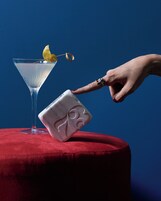 Une main qui incline un objet blanc orné d’un logo sur une surface rouge, à côté d’un verre transparent décoré d’une tranche de citron.