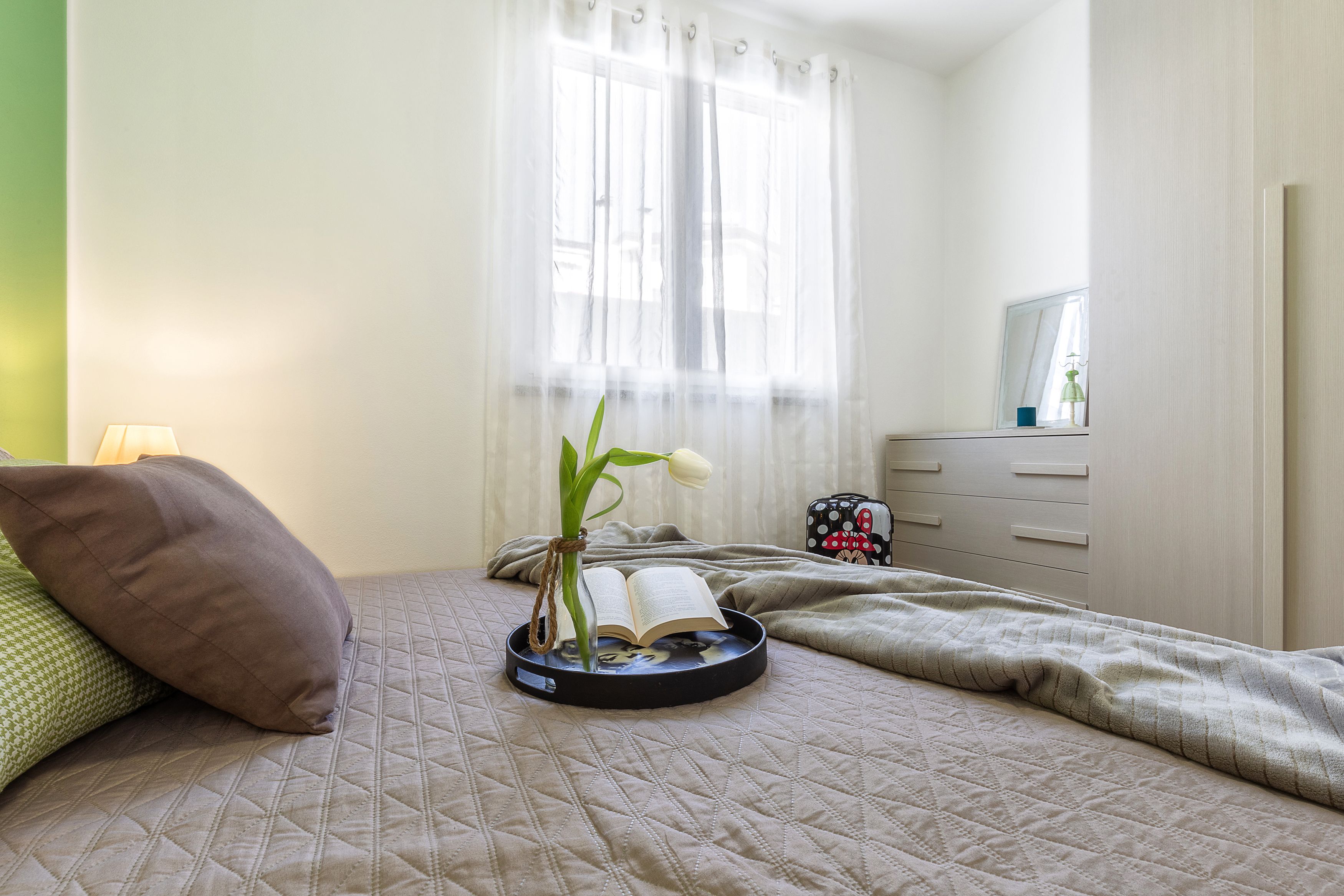A neatly made bed with a quilted cover, a bedside table with a book and a plant, and a window with sheer curtains.