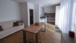 A modern living room with a wooden dining table, a sofa, and a kitchen in the background. - Casa Selvaggio Blu (Baunei)