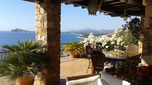 A terrace with stone pillars, a table with chairs, potted plants, and a view of the sea and mountains. - Casa sul Porto (Baunei)