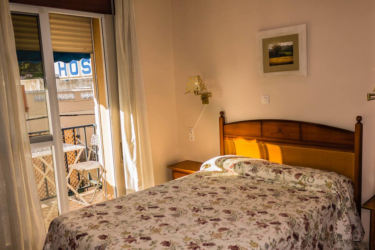 Ein Schlafzimmer mit Bett, Nachttisch, Lampe, einem Bild an der Wand und Blick auf den Balkon.