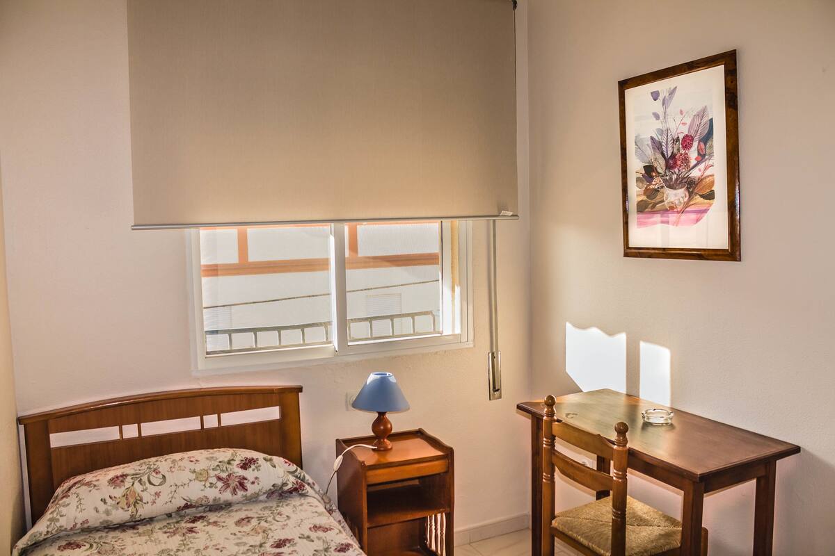 Ein Schlafzimmer mit Bett, Nachttisch mit Lampe, einem Holzstuhl, einem kleinen Tisch und einem Fenster mit Blick.