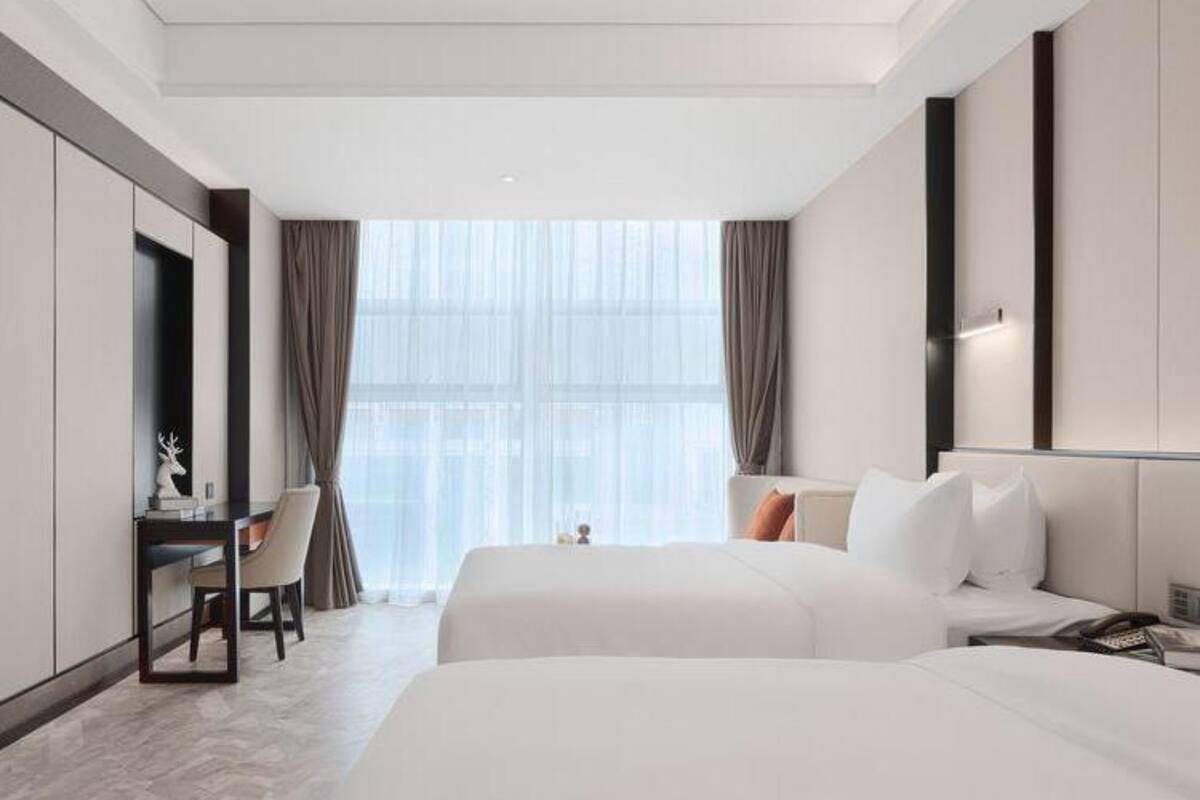 Ein modernes Hotelzimmer mit zwei Betten, einem Schreibtisch und einem großen Fenster mit Vorhängen.