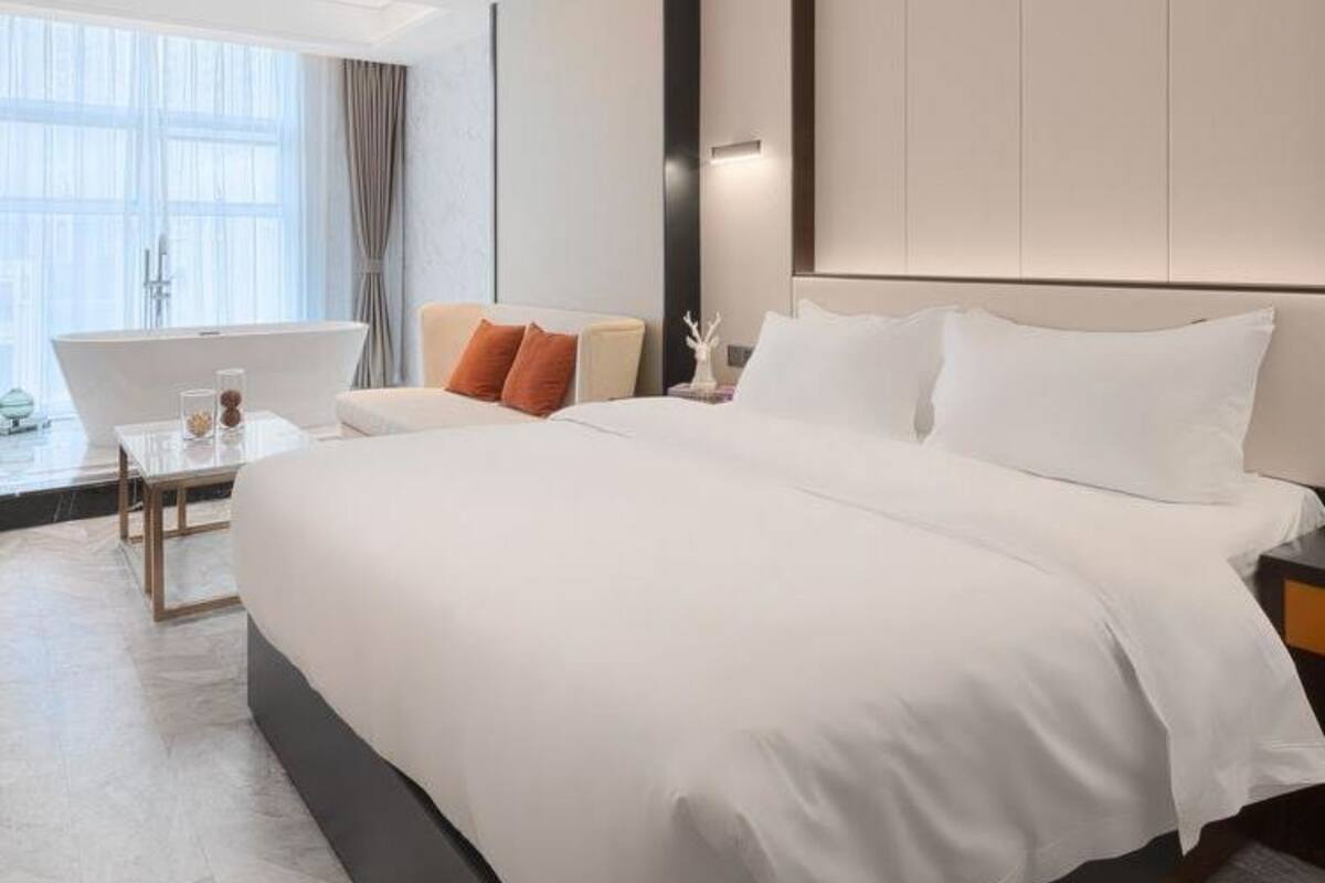 Ein modernes Hotelzimmer mit einem großen Bett, einer Sitzecke mit einer Couch und Sesseln, einem kleinen Tisch und einem großen Fenster mit Vorhängen.
