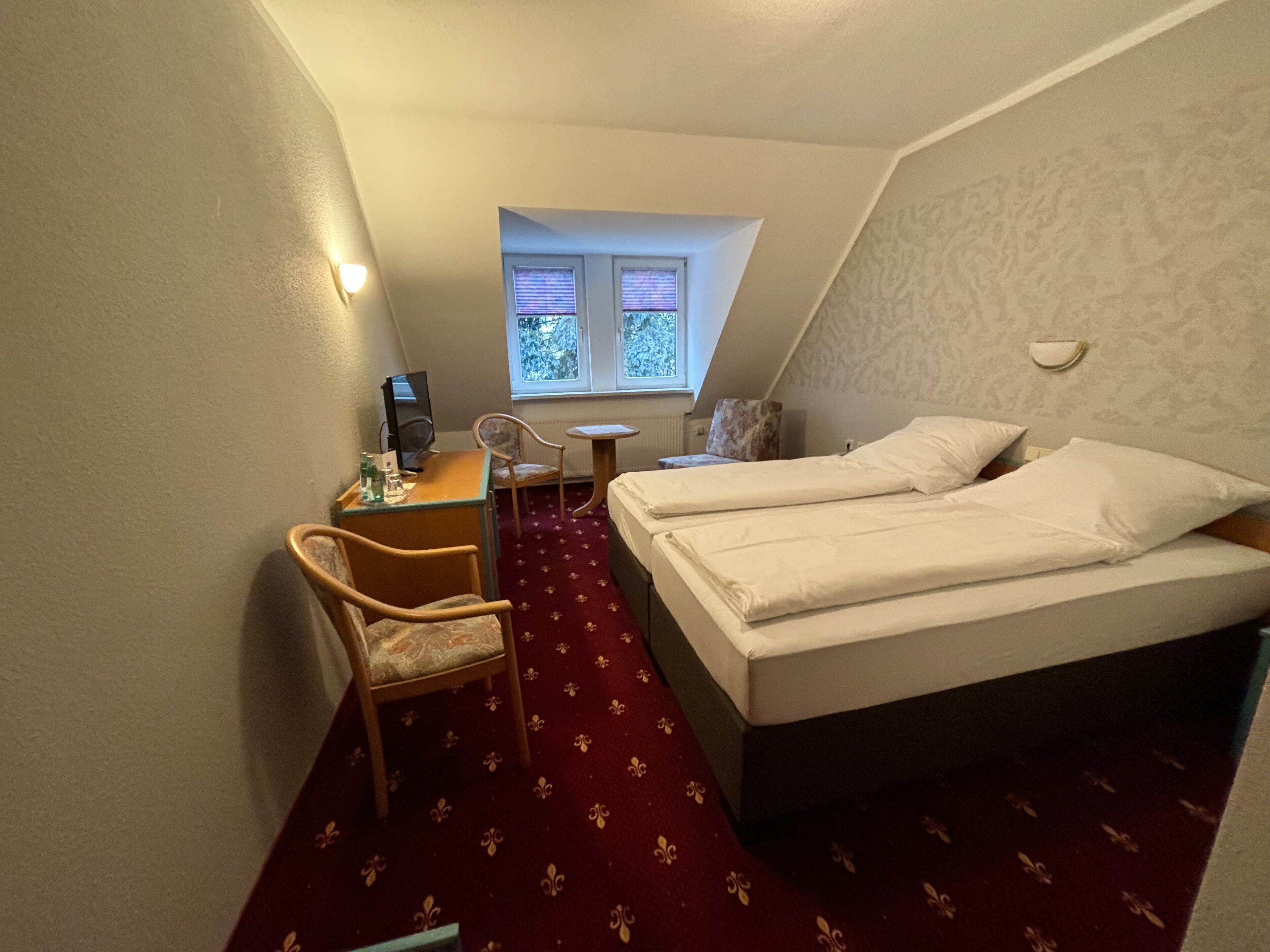 Um quarto pequeno e triangular com uma cama, uma escrivaninha e uma cadeira. Há uma janela com persianas, um radiador e uma luminária fixada na parede.