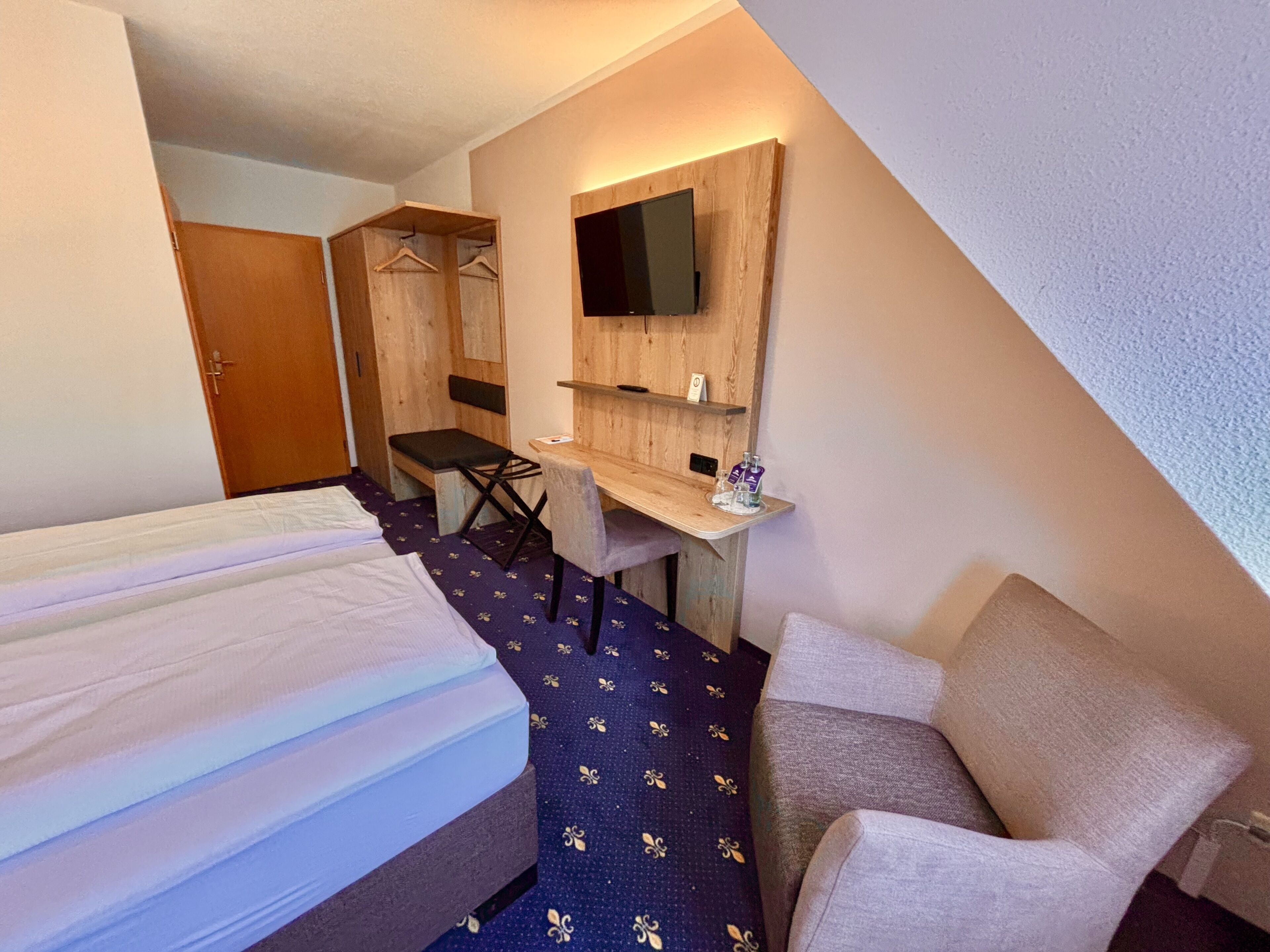 Habitación de hotel con cama, escritorio, silla, televisor montado en la pared y armario.