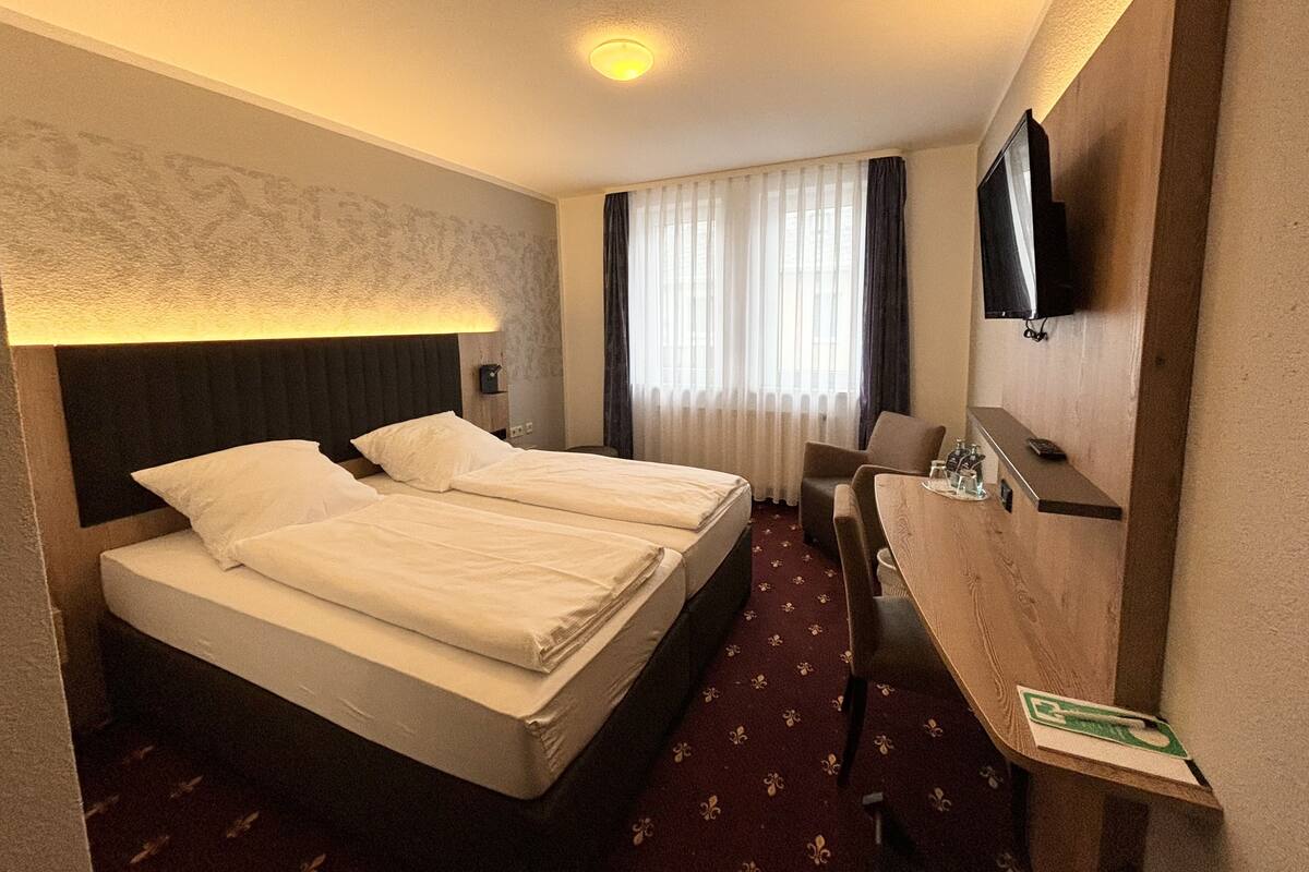 A hotel room with two single beds, a desk with a chair, a flat-screen TV mounted on the wall, and a window with sheer curtains.