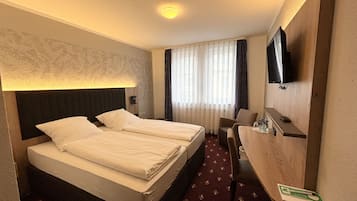 Ein Hotelzimmer mit zwei Einzelbetten, einem Schreibtisch mit Stuhl, einem flachbildfernseher an der Wand und einem Fenster mit durchsichtigen Vorhängen.