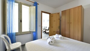 A hotel room with a bed, a wooden wardrobe, a white chair, and a window with blue curtains. - Casa Cala Sisine (Baunei)