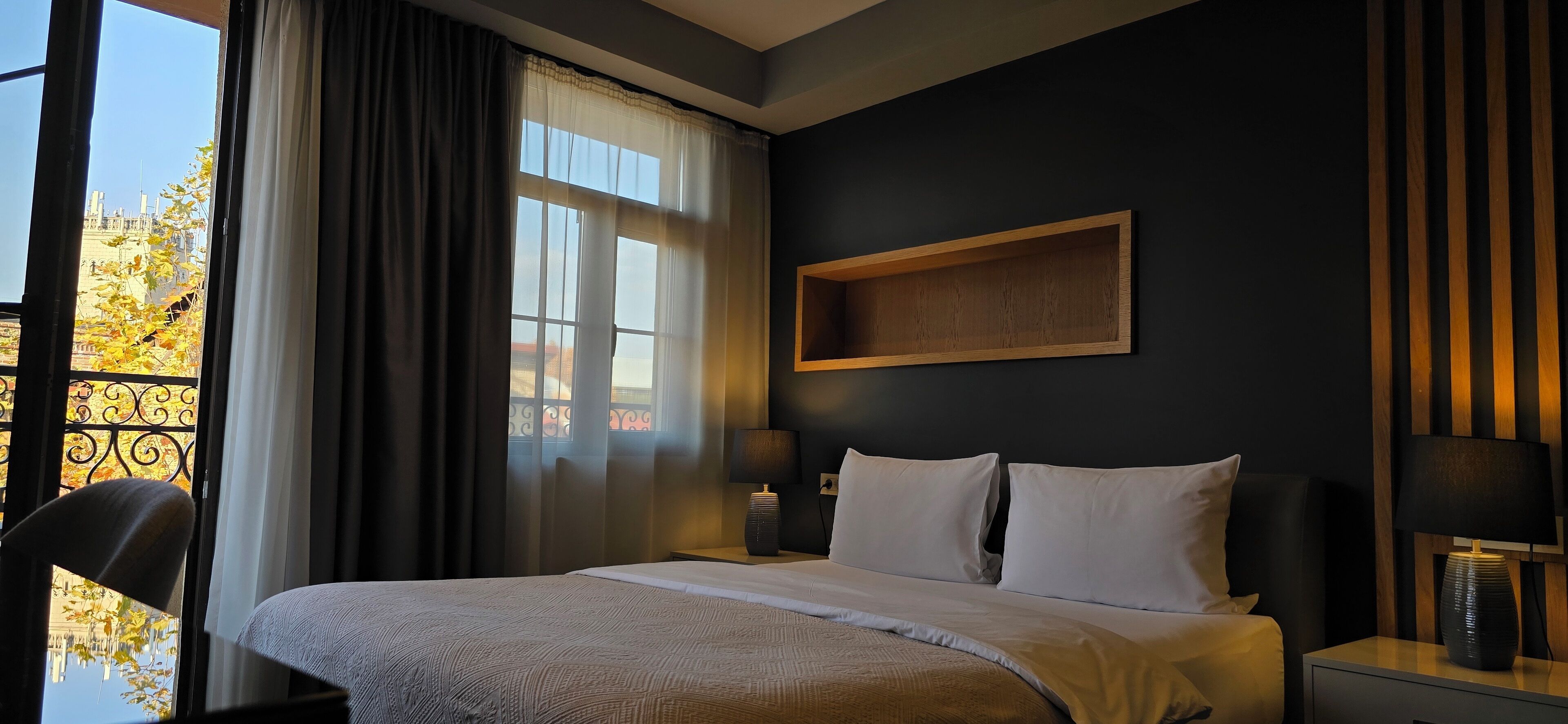 A hotel room with a large bed, two bedside tables with lamps, a dark wall, and a window with curtains.