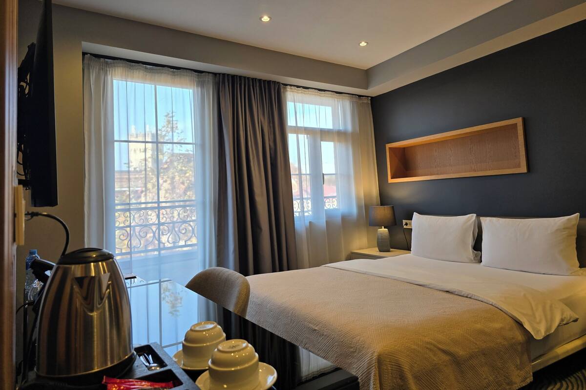 A hotel room with a bed, a TV, a window with curtains, a bedside table with a lamp, and a wall-mounted shelf.