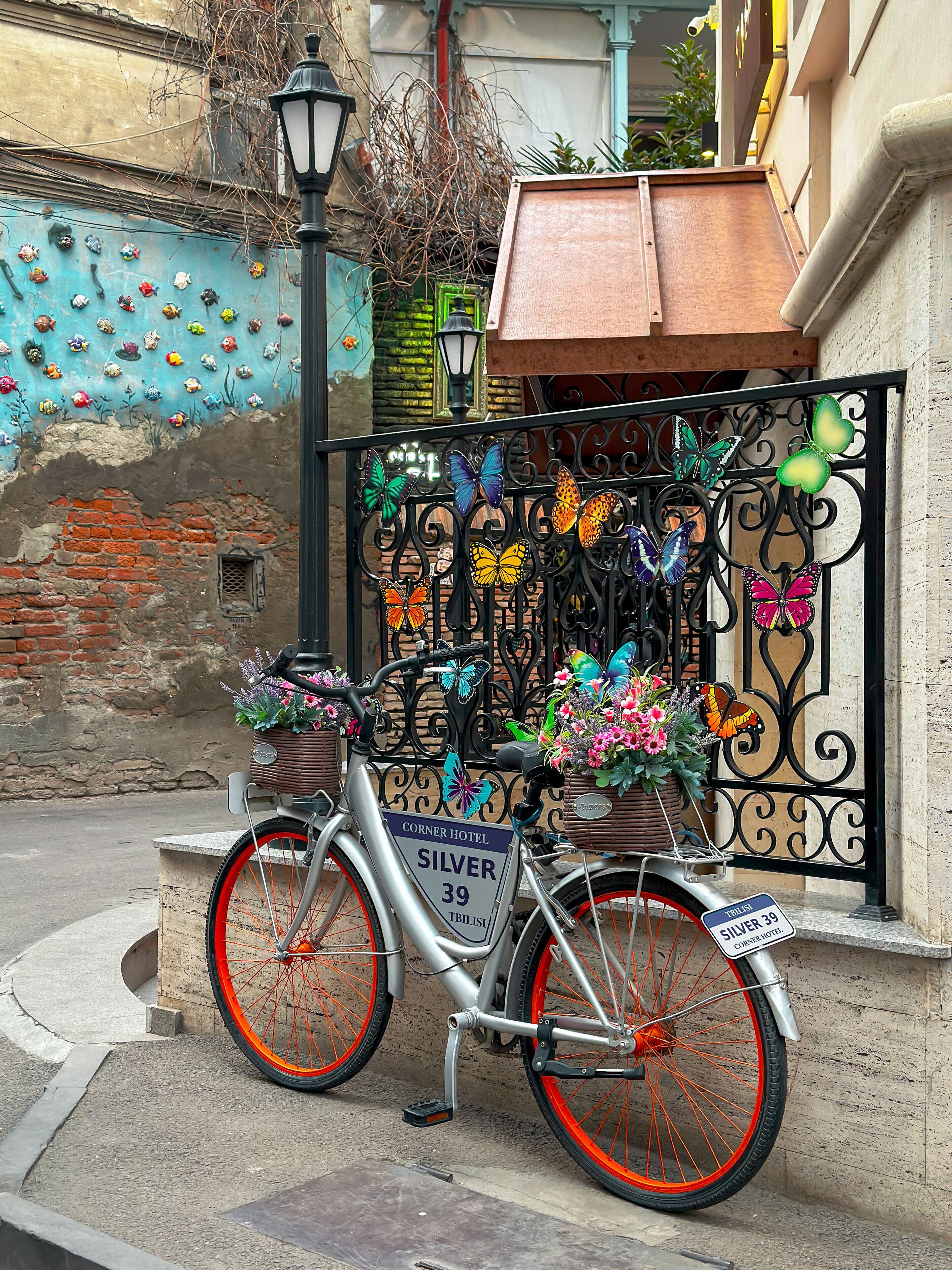 Ein Fahrrad vor einem Gebäude mit einem verzierten Tor, das Schmetterlinge zeigt. Auf dem Schild des Fahrrads steht "CORNER HOTEL SILVER 39".