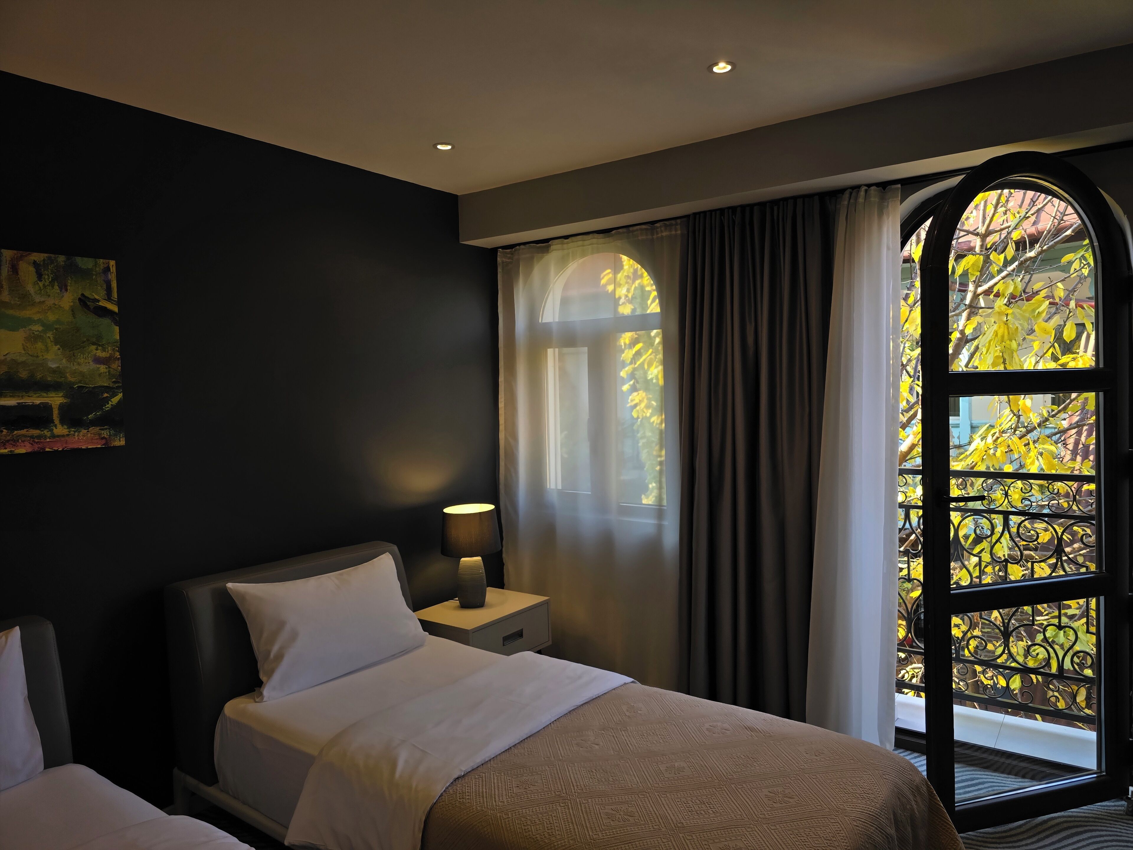 Ein Hotelzimmer mit Bett, Nachttisch, Lampe und einem großen Fenster mit Vorhängen.