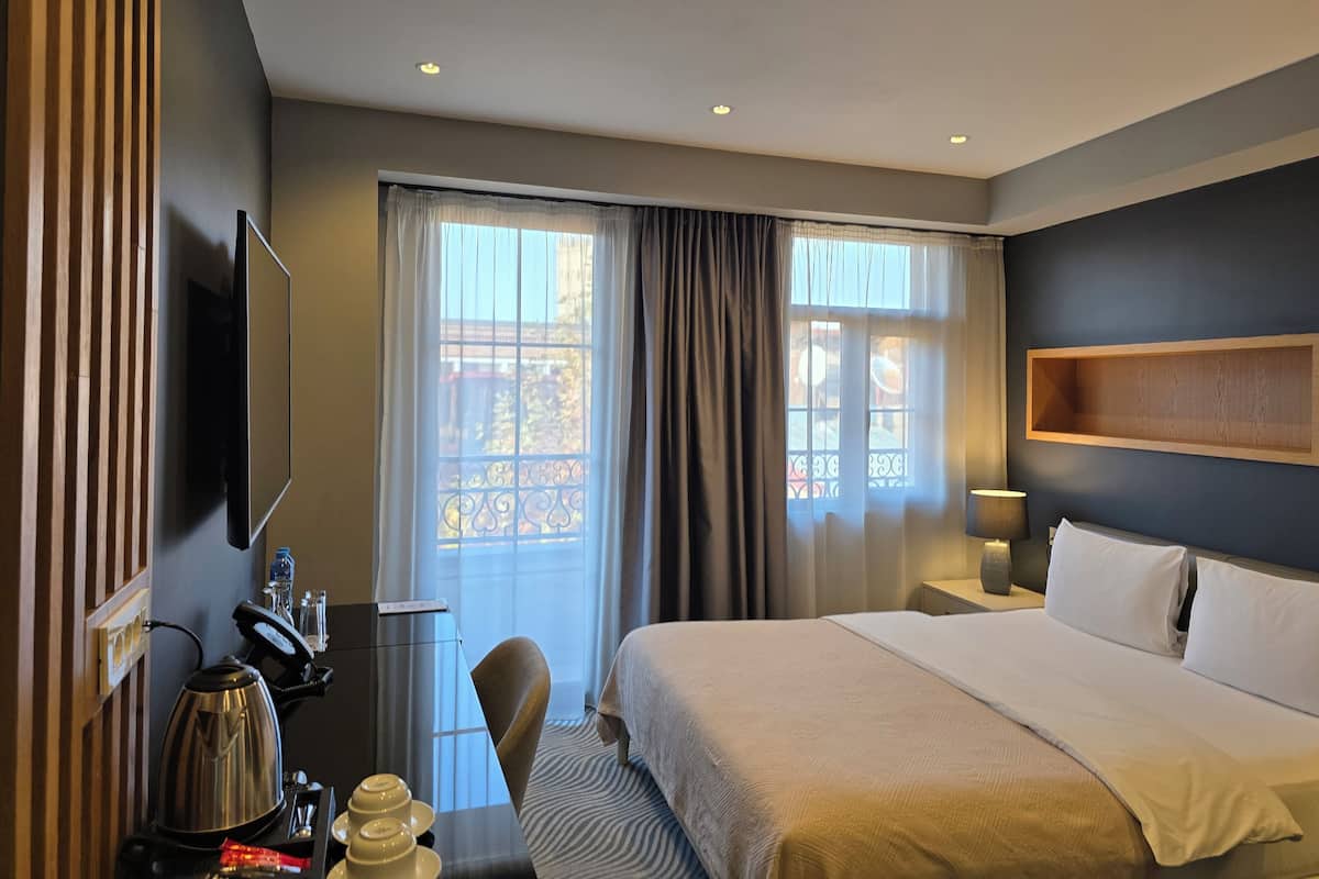 A hotel room with a bed, a desk with a kettle, a chair, a TV, and a window with curtains.