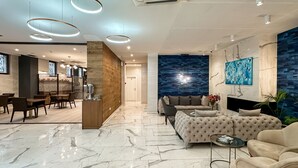 Een moderne hotellobby met een marmeren vloer, een zithoek met banken en fauteuils, een bar en een groot abstract schilderij in blauwe tinten aan de wand.