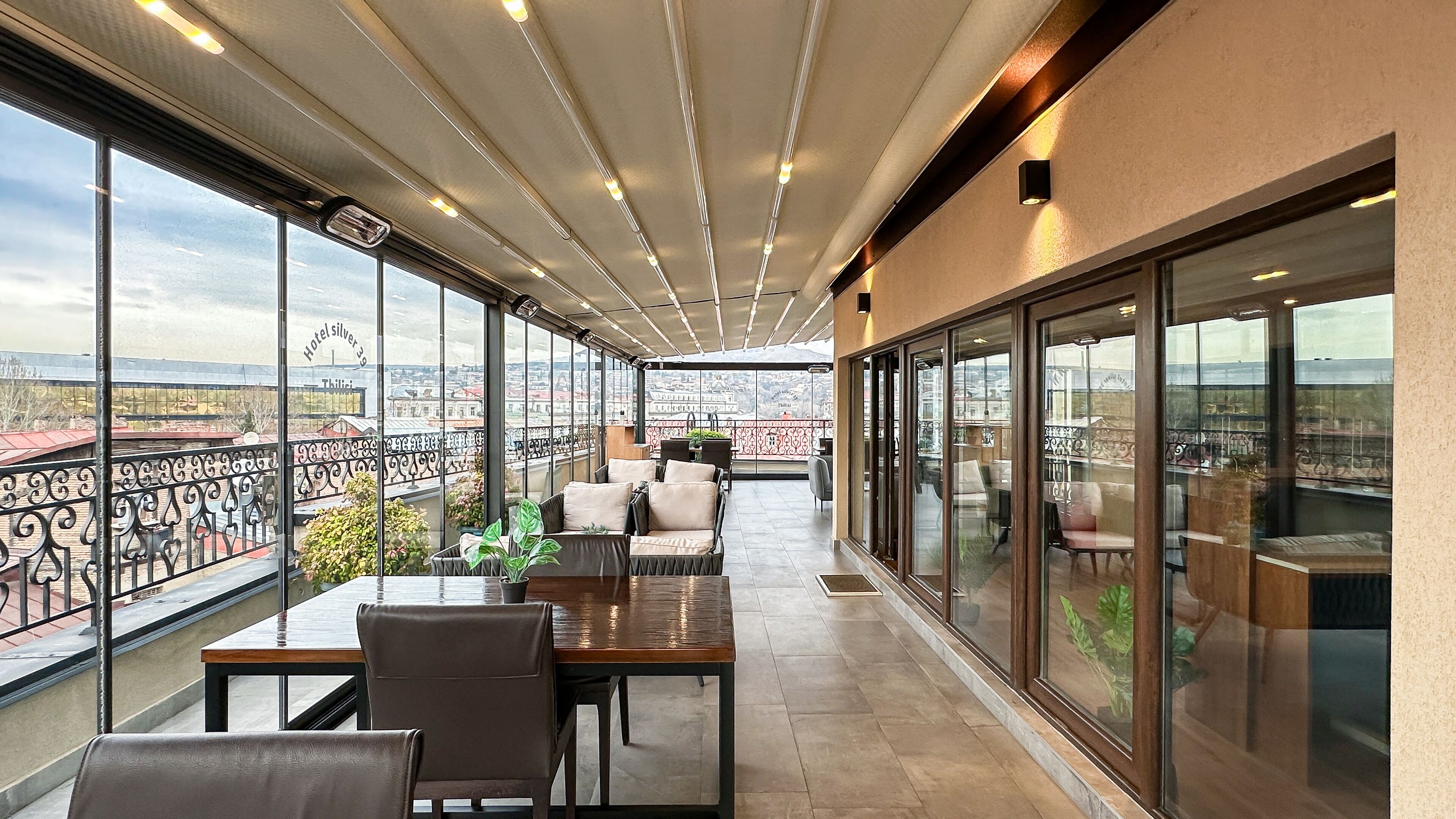 En rummelig balkon med bord og stole, store vinduer der giver udsigt over byen, og et loft med lineære lysarmaturer.