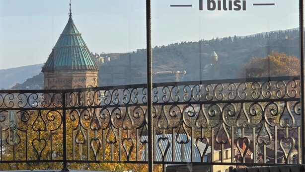 Een balkon met een tafel gedekt voor twee personen, compleet met wijnglazen en een vaas met gele bloemen. Een sierhek van metaal en een toren in de verte zijn ook te zien.