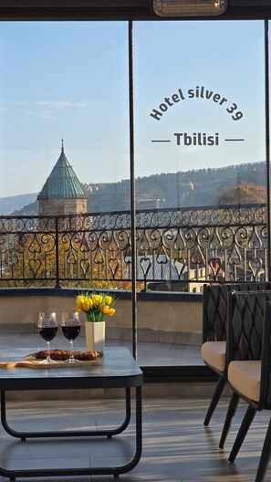 A balcony with a table set for two, featuring wine glasses and a vase of yellow flowers. A decorative metal fence and a view of a tower in the background are also visible. - Silver 39 Corner Hotel (Tbilisi)