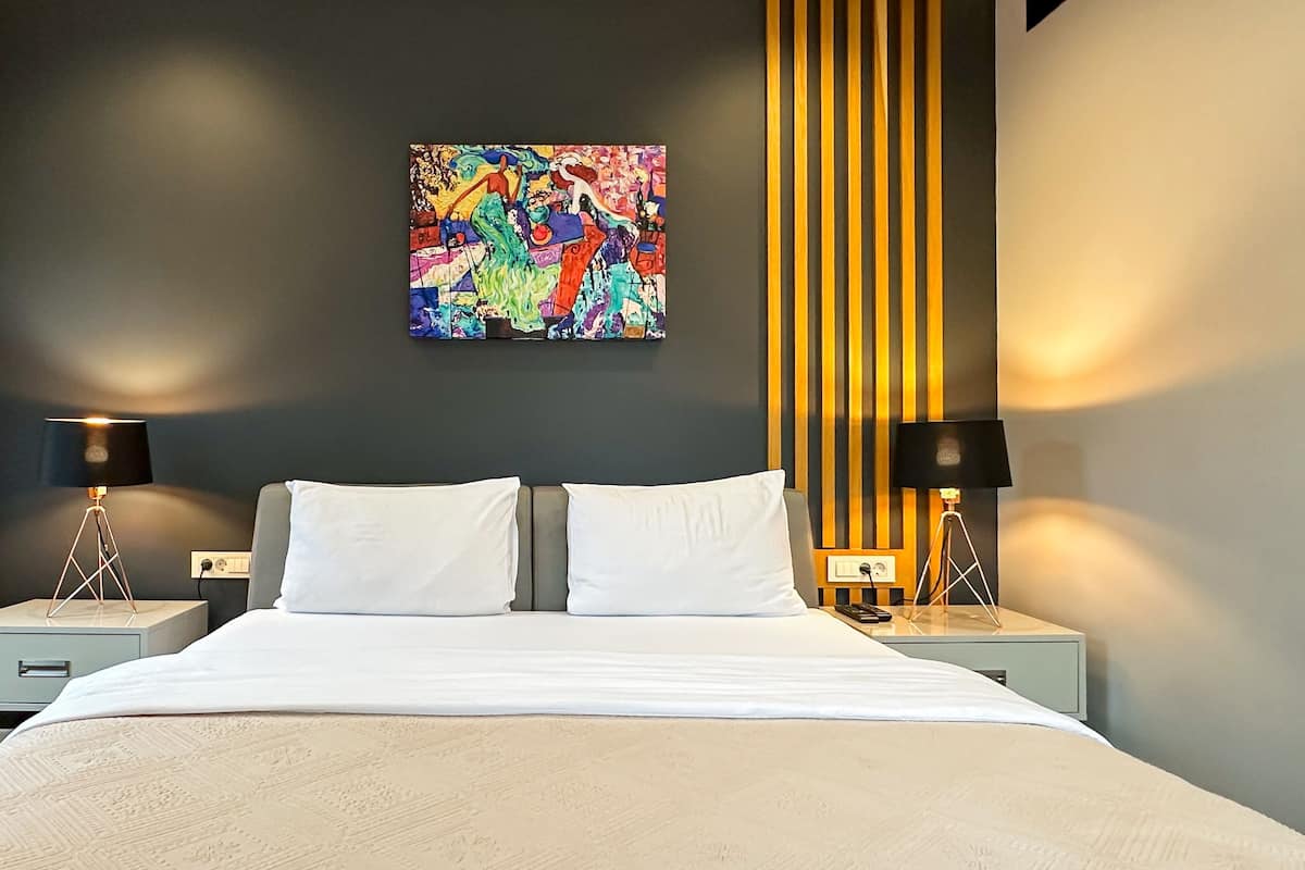A modern hotel room with a large bed, two bedside tables with lamps, a colorful abstract painting on the wall, and a vertical light panel.