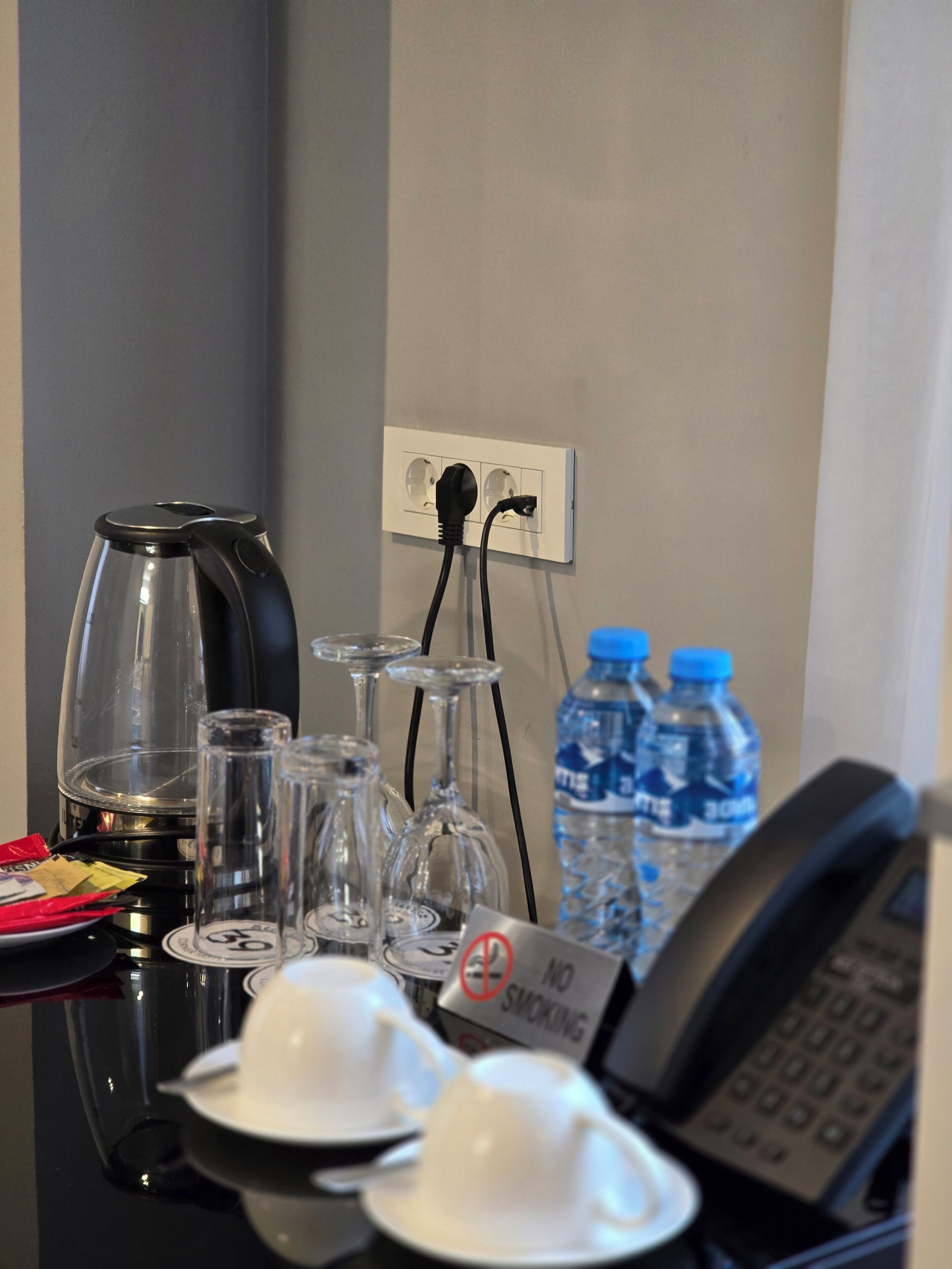 Een bureau in een hotelkamer met een waterkoker, glazen, een waterfles, een rookverbodsgevaarsbord en een telefoon.