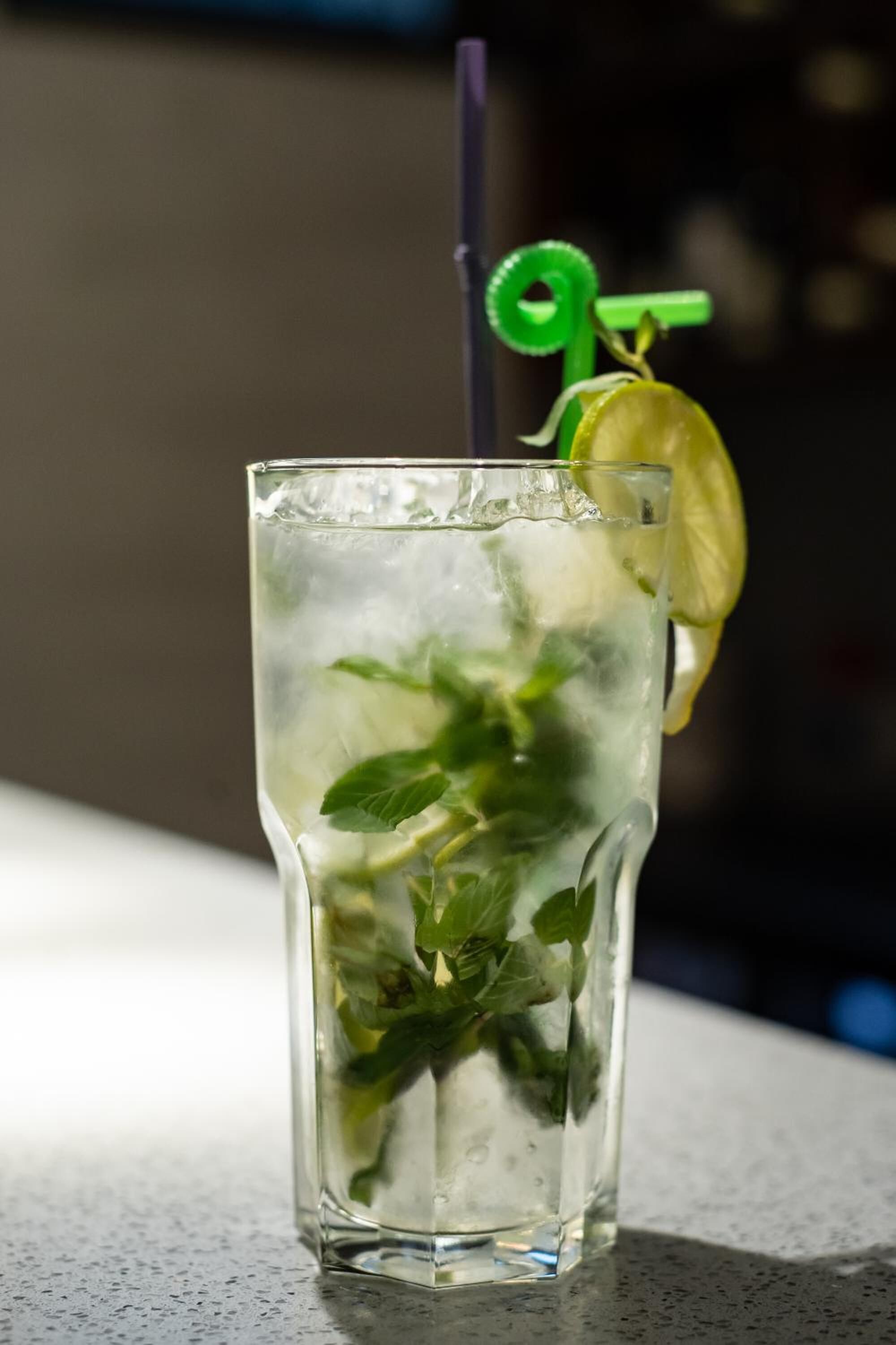 A tall glass with ice, mint leaves, and a lemon slice, with a green straw and a green decorative item.