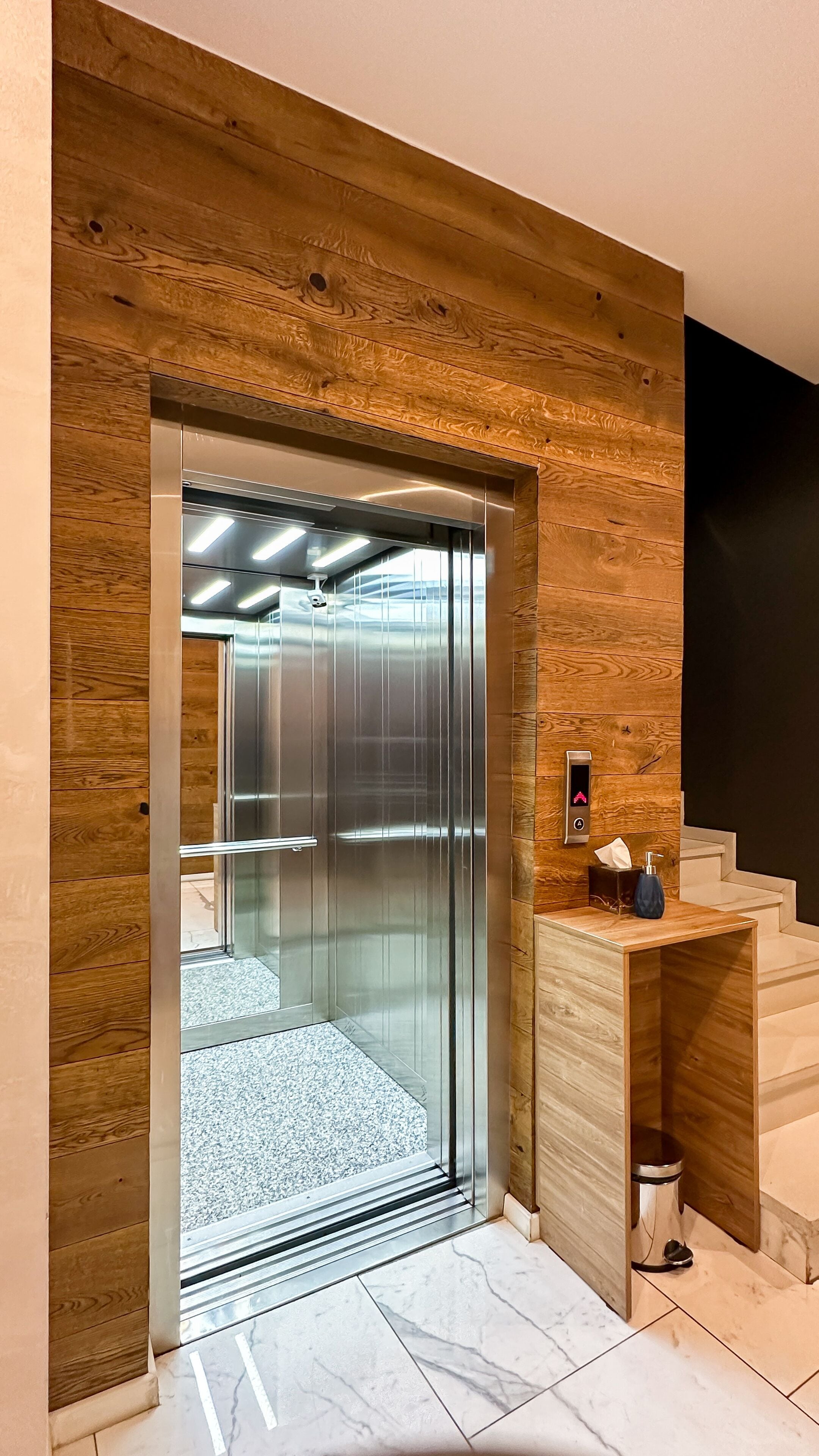 A modern elevator with a wooden panel design, a visible emergency phone, and a marble-tiled floor.