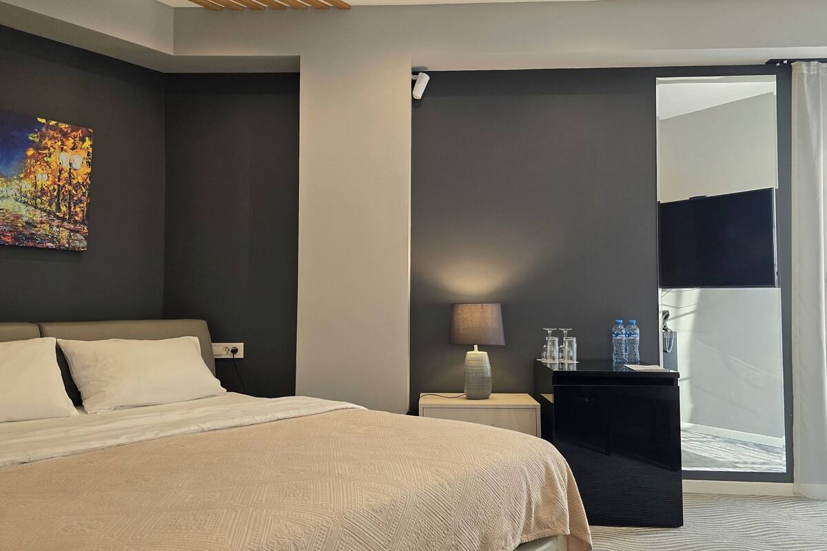 A hotel room with a bed, a nightstand, a lamp, a painting, and a view of the outside.